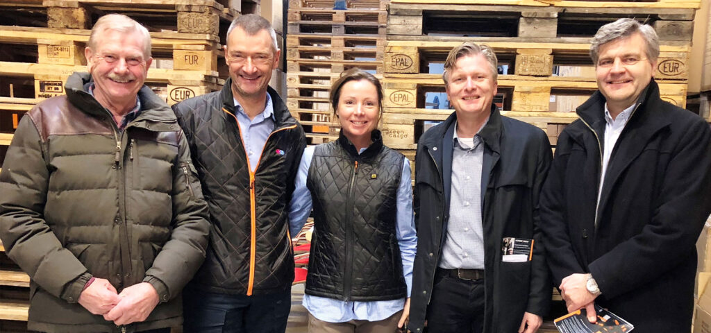 Žmonės dėvi dygsniuotus liemenes priešais sukrautus padėklus. NordicHeat produktai laikomi elegantiškais šiltais drabužiais, tinkamais aktyviam gyvenimo būdui. Tekstas: "NORDIC HEAT" ant striukės kišenės klosčių.