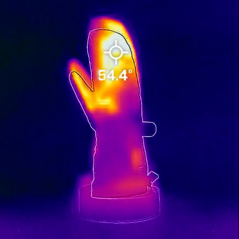 Termovizinis vaizdas rodo kumštinę pirštinę iš NordicHeat su matoma temperatūros indikacija 54.4 laipsnių. Kumštinė pirštinė atrodo šilta centre, o aplinka lieka šalta.