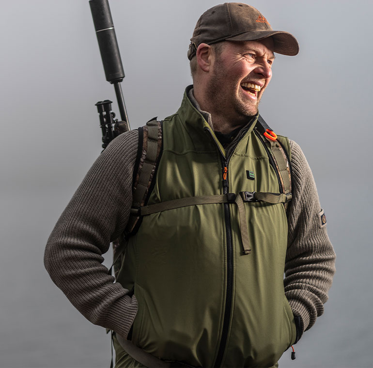 A smiling person wearing NordicHeat warm clothes and a cap stands outdoors with a backpack and a rifle strapped to his back. The atmosphere seems humid or foggy.