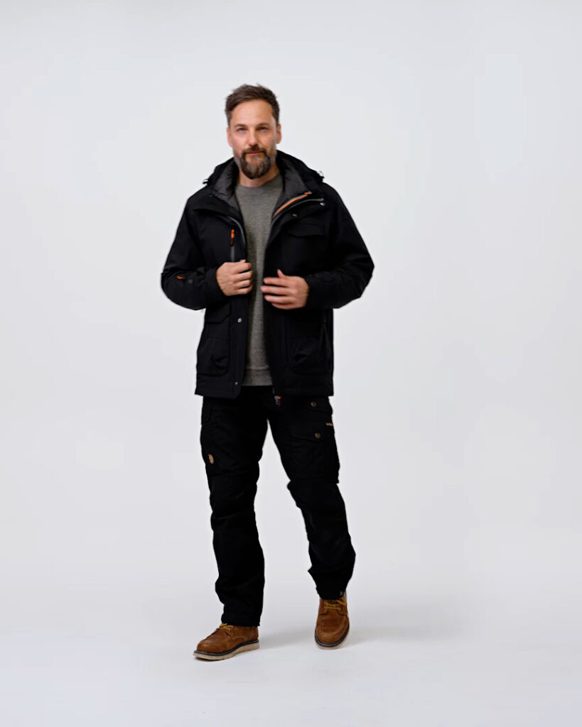Vyras, dėvintis juodus NordicHeat termo drabužius, stovi neutralioje studijos aplinkoje. Jis turi rusvus batus, pilkus marškinius ir tvarkingą barzdą, su ramiu kūno kalba į kamerą.