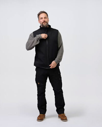 A man wearing NordicHeat thermal clothing adjusts the zipper on a black vest. He is standing in a neutral studio with casual clothing, which includes pants and brown boots.