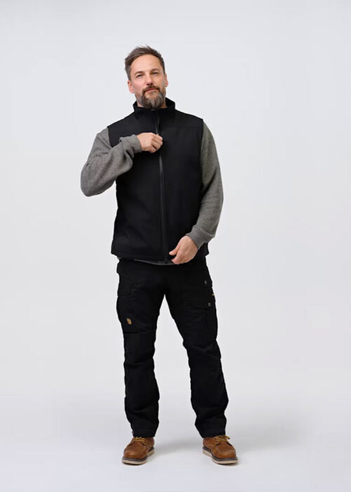 A man wearing NordicHeat thermal clothing adjusts the zipper on a black vest. He is standing in a neutral studio with casual clothing, which includes pants and brown boots.