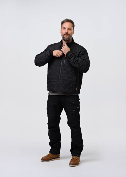 A person wearing a black quilted jacket and pants stands against a white background, adjusting the jacket at the neck. The person has brown hair and a beard.