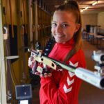 A person in a shooting range wearing a red sports jacket with white arrow patterns, aiming a rifle, highlighting NordicHeat's warm clothing for active lifestyle. No visible text.