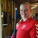 Object: Person in red jacket with text "Danish Shooting Team" and weapon; Action: Smiling at shooting station; Context: Indoor shooting range, representing active lifestyle and performance.