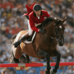 Rider wearing red jacket and helmet jumps over obstacle on horse in sports event. No visible text. NordicHeat warm clothing can optimize comfort and performance during such active outdoor activities.