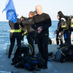 Diver dressed in NordicHeat warm clothing prepares at sea with diving equipment. Text: "Nordic Heat." Active lifestyle with a focus on keeping warm outdoors.
