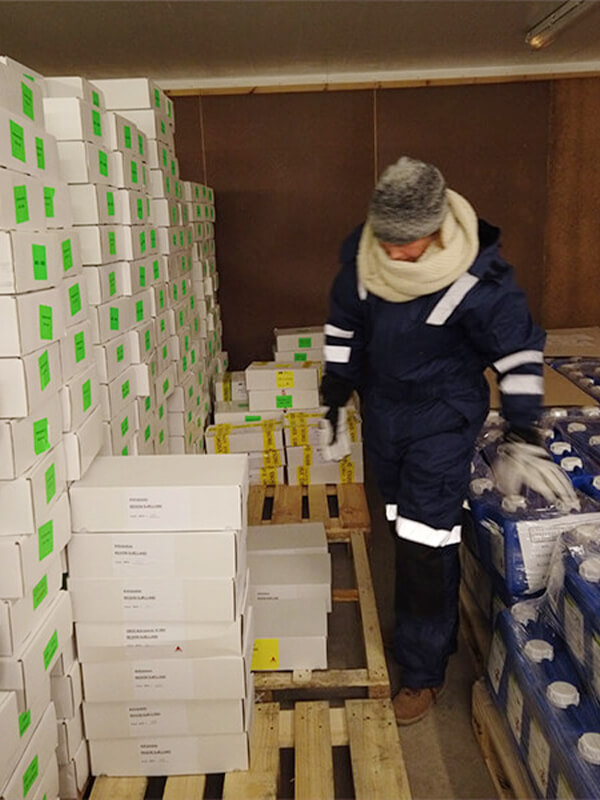 Asmuo, dėvintis NordicHeat šiltus drabužius, juda tarp sukrautų dėžių šaltoje patalpoje, dirbdamas sandėlio darbus. Šilumos izoliacijos produktai leidžia aktyvų gyvenimo būdą šaltose aplinkose. Matomas tekstas: [nėra matomo teksto].