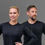 Two people wearing black NordicHeat thermal clothing stand in front of a gray background. The clothes have a visible zipper at the neck.