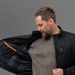 A man wearing a gray sweater opens a black NordicHeat jacket, revealing an orange edged inner pocket. The background is solid gray.