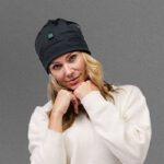 A woman wearing a white sweater and a black beanie from NordicHeat stands in front of a gray background, hands raised to her chin.