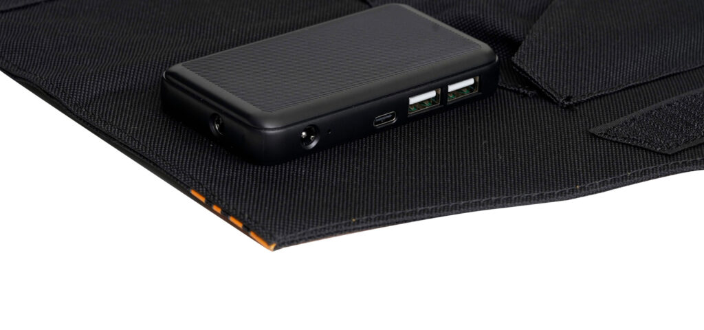 Nešiojama baterija padėta ant juodos tekstilės medžiagos. Baterija turi USB išvestis, nurodo naudojimą NordicHeat šiluminėje aprangoje, palaiko aktyvų lauko gyvenimo būdą. Matomas tekstas: Nėra.
