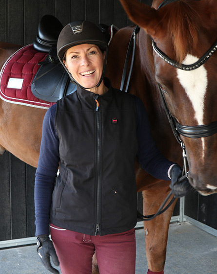 A person wearing a black NordicHeat vest is standing next to a horse, wearing a riding helmet and gloves, in a stable situation.