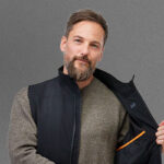 A man shows an open black heated vest from NordicHeat, wearing a gray sweater, against a gray background.