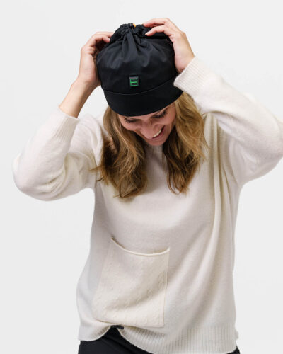 Person adjusts black NordicHeat beanie with heating panel, dressed in white sweater, symbolizing active lifestyle and practical winter clothing.