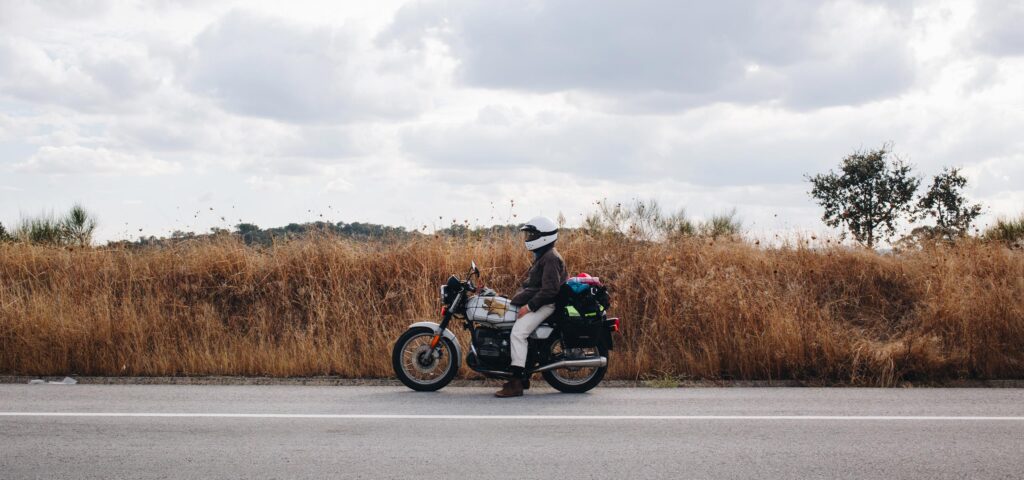 Motociklininkas, dėvintis NordicHeat šalmą ir striukę, sustoja kelkraštyje, apsuptas gamtos, simbolizuojantis aktyvų gyvenimo būdą su efektyviais termo drabužių deriniais. Nėra matomo teksto.